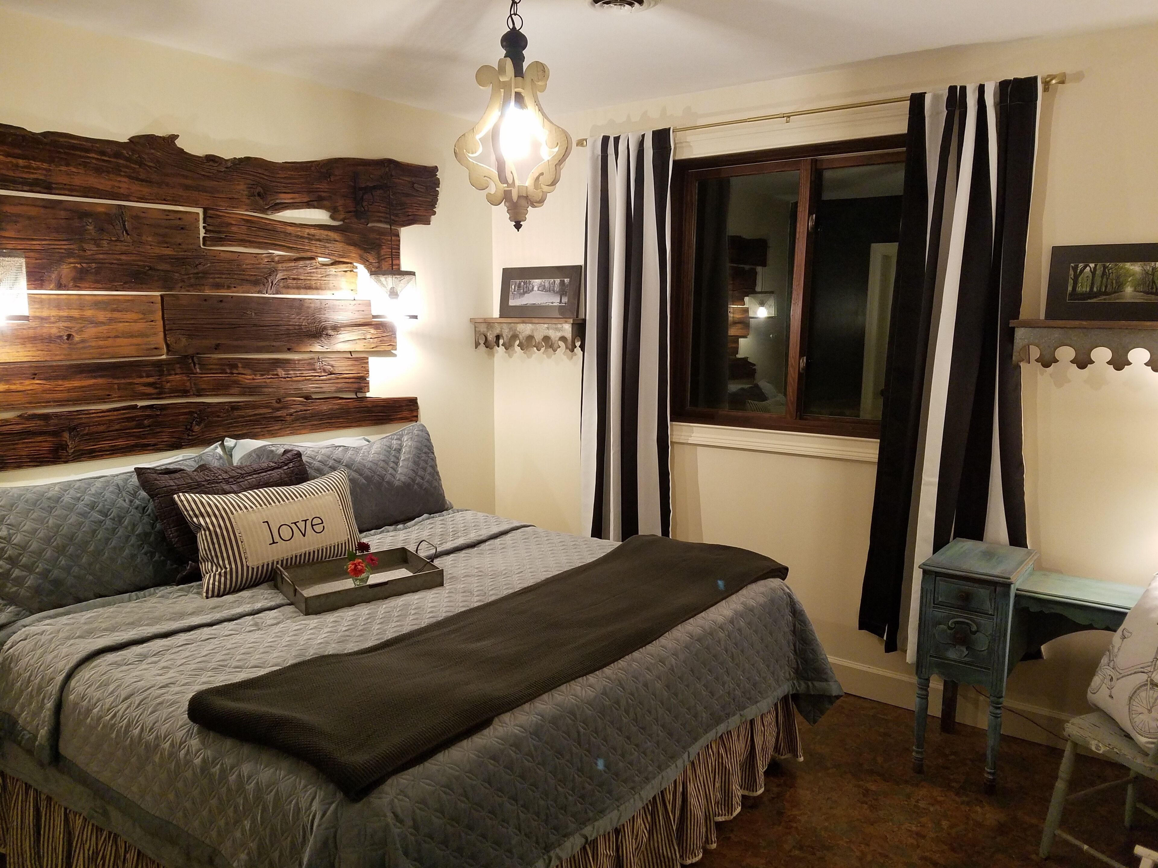 Un dormitorio con cabecera de madera, cama con colcha acolchada, mesita de noche con lámpara, ventana con cortinas y una silla pequeña.