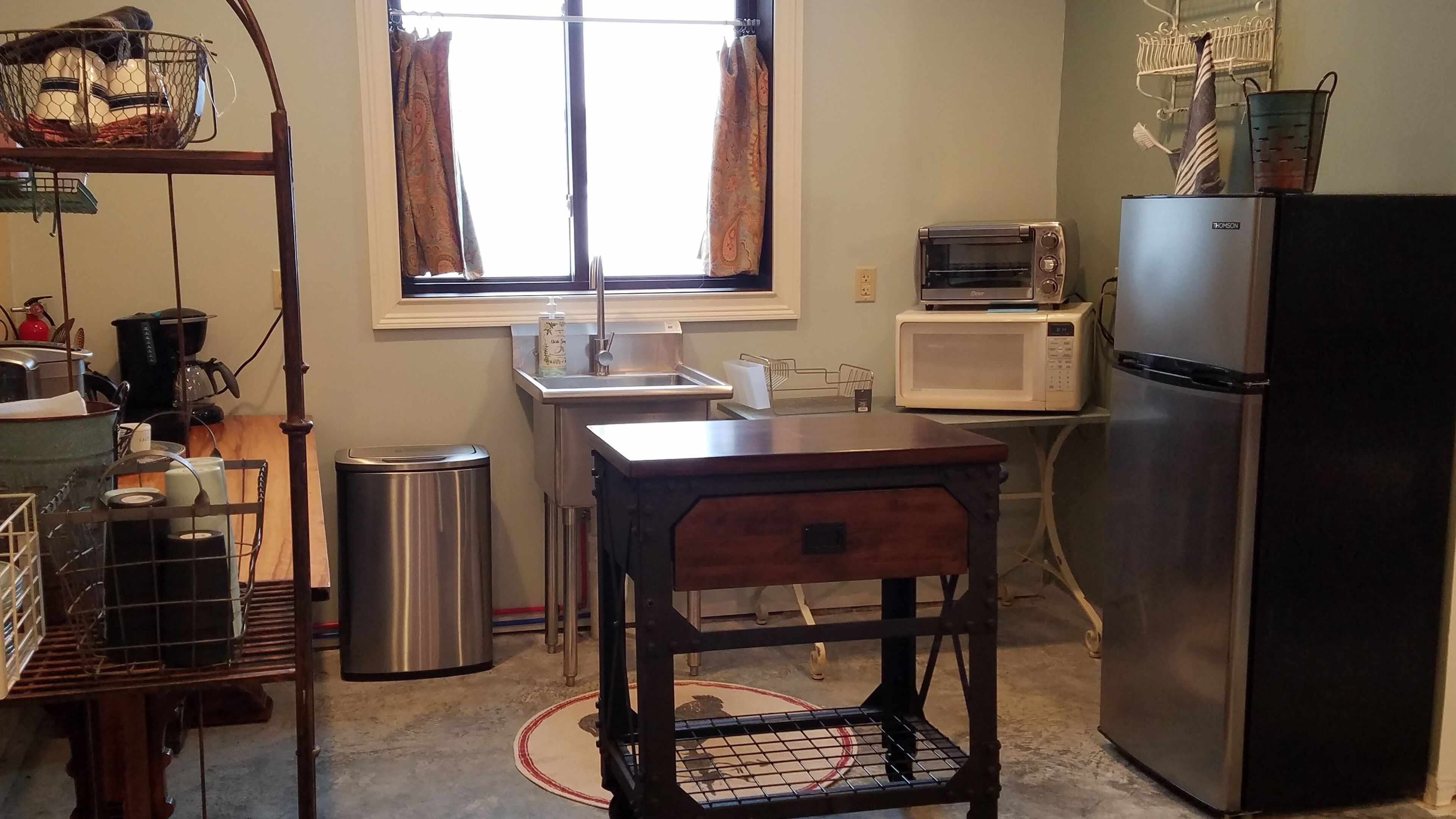 Una cocina compacta con un cubo de basura de acero inoxidable, una mesa de madera, un microondas, un tostador y un refrigerador.