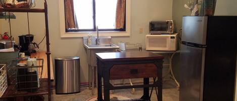Una cocina compacta con un cubo de basura de acero inoxidable, una mesa de madera, un microondas, un tostador y un refrigerador.