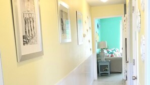 A hallway with framed pictures on the walls, a bed with a green wall in the background, and a door on the right.
