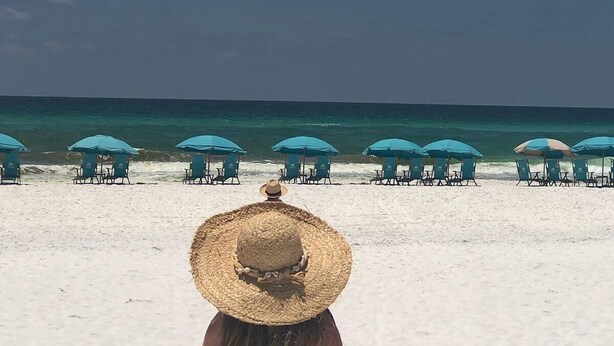 A beach with rows of blue umbrellas and chairs, a clear sky, and a person in the foreground wearing a straw hat.