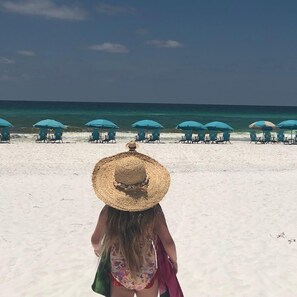 A beach with rows of blue umbrellas and chairs, a clear sky, and a person in the foreground wearing a straw hat.