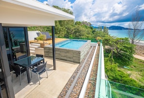 Un área de piscina exterior moderna con una mesa y sillas de vidrio, rodeada por una terraza de piedra y vistas al mar y a las colinas.