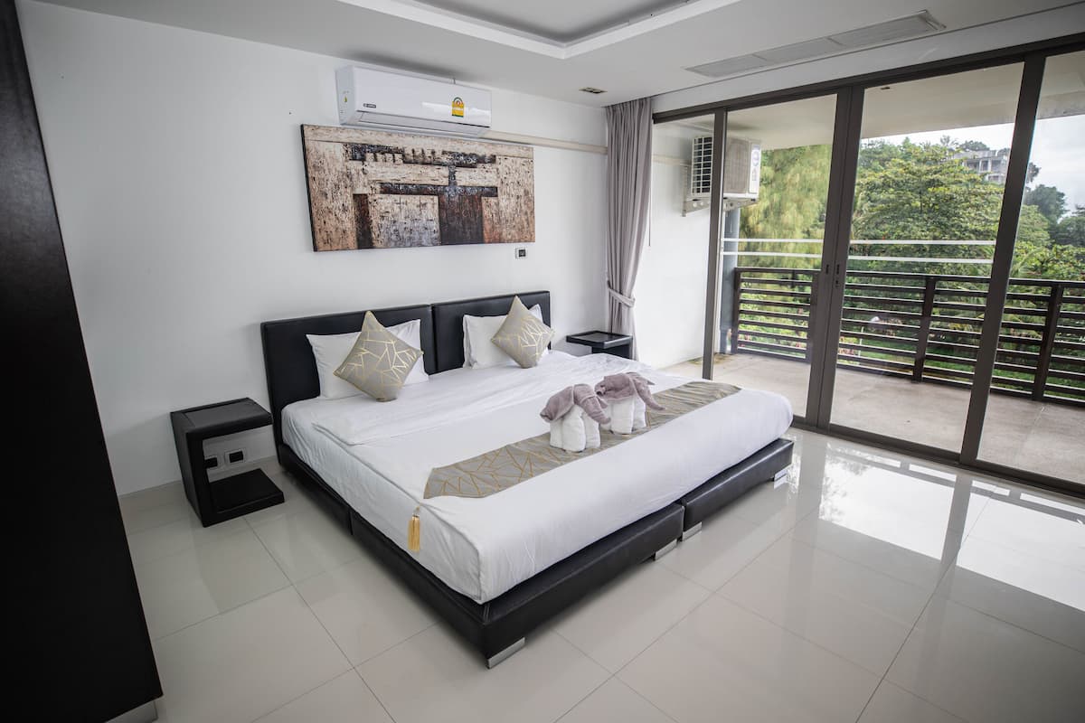 A modern bedroom with a large bed, a nightstand, a wall-mounted air conditioner, and a view of a balcony with greenery.