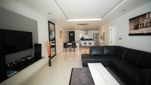 A modern living room with a flat-screen TV, black leather sofa, and a dining area with a kitchen in the background. - Nakalay Seaview Penthouse (Kamala)
