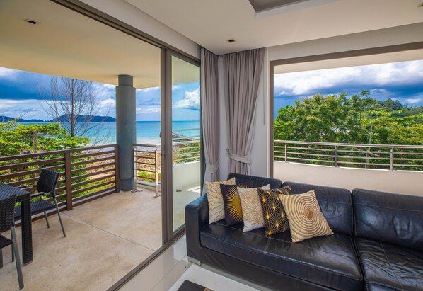 A balcony with a view of the sea and mountains, a black leather sofa with patterned cushions, and a dining area with chairs.