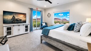 Quarto com cama, TV, ventilador de teto, janela vista para o deserto e varanda com cadeira.