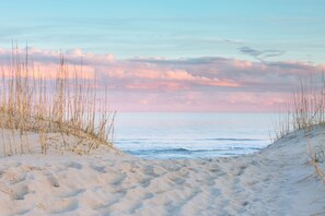 Una playa arenosa con hierbas altas y un mar tranquilo bajo un cielo rosado y azul.