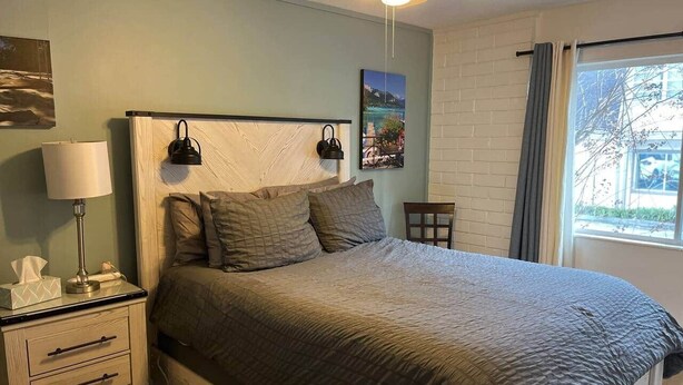 Un dormitorio con cama, mesita de noche, lámpara y ventana con cortinas.