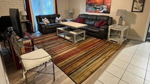 Una sala de estar con un sofá de cuero negro, un sillón blanco, una mesa de centro de madera y una alfombra a rayas.