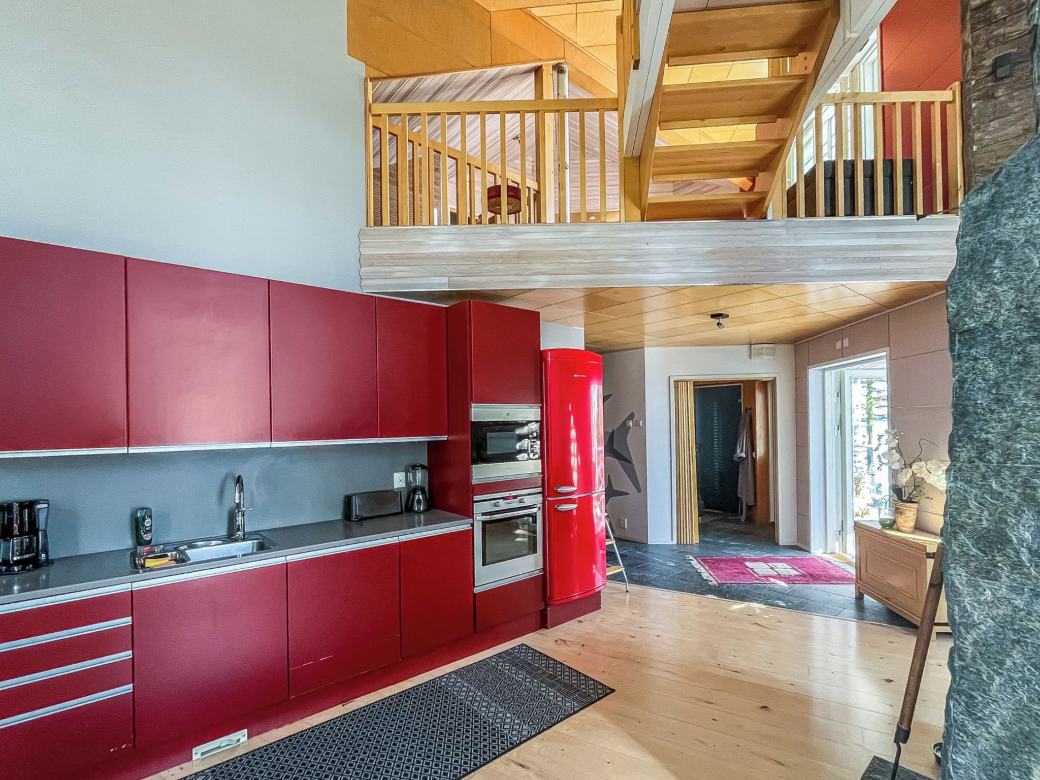 A modern kitchen with red cabinets, a red refrigerator, and a wooden staircase leading to an upper level.