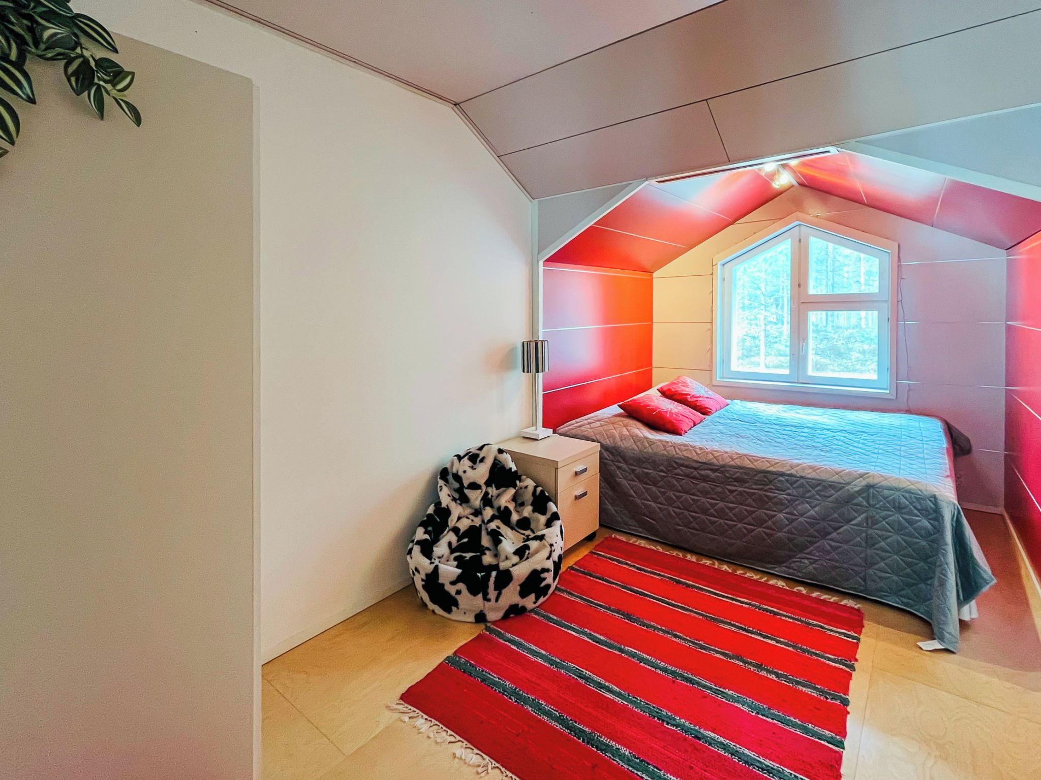 Ein Schlafzimmer mit schrägen Wänden, einem Bett mit gesteppter Bettdecke, einem roten und schwarzen Sitzsack sowie einem Fenster mit Blick auf Bäume.