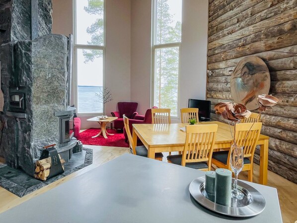 A cozy living area with a stone fireplace, a dining table with chairs, a red armchair, and a view of the outdoors through large windows.