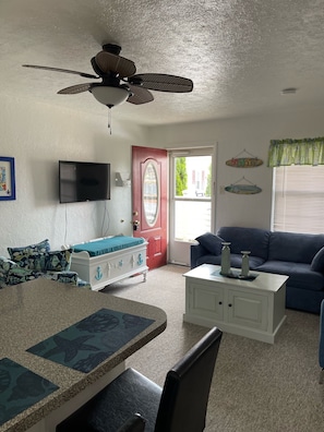 A living room with a ceiling fan, a red door, a television, a blue couch, and a dining area with a table and chairs. - LUXURY 2 BEDROOM 1ST FLOOR CONDO WITH A POOL (Wildwood)