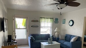 A living room with a blue sofa, a white coffee table, and a ceiling fan. There is a window with curtains, a wall clock, and a television mounted on the wall.