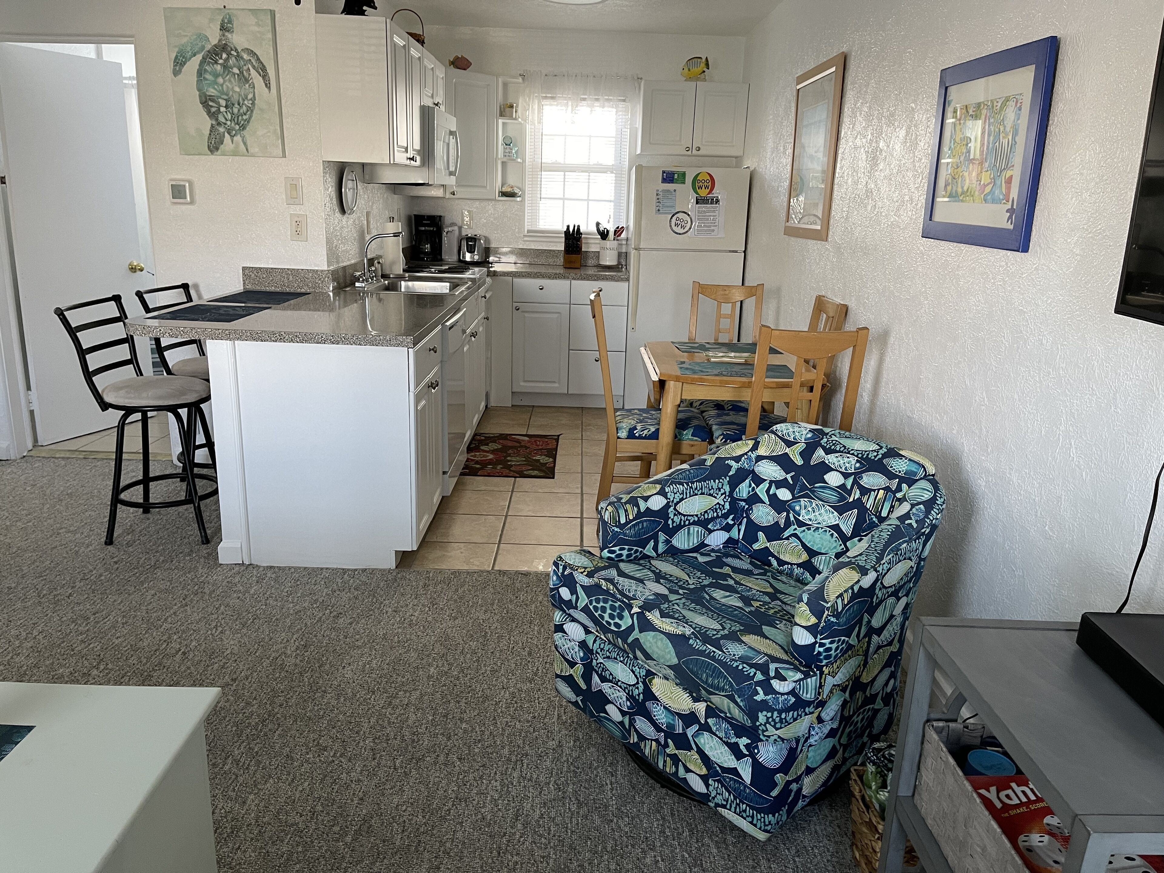 Een kleine keuken met witte kasten, een koelkast en een eethoek met een gestreept fauteuil en houten stoelen.