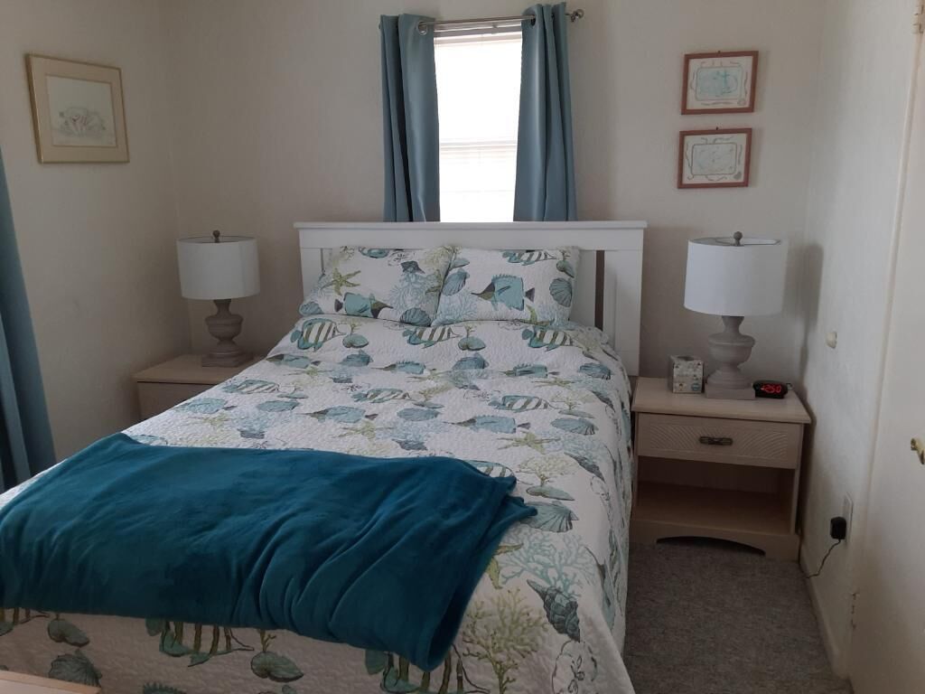 Un dormitorio con una cama, dos mesitas de noche, una mesita auxiliar, una ventana con cortinas y cuadros en la pared.