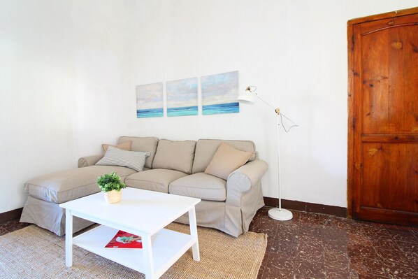 Una sala de estar con un sofá color beige, una mesa de centro blanca, una planta pequeña y un cuadro con una playa.