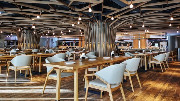 Un restaurant moderne avec des tables et des chaises en bois, un plafond haut avec des poutres apparentes et des luminaires suspendus.