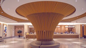 Un hall d’hôtel moderne avec une colonne centrale en bois, un comptoir d’accueil et une grande plante en pot.