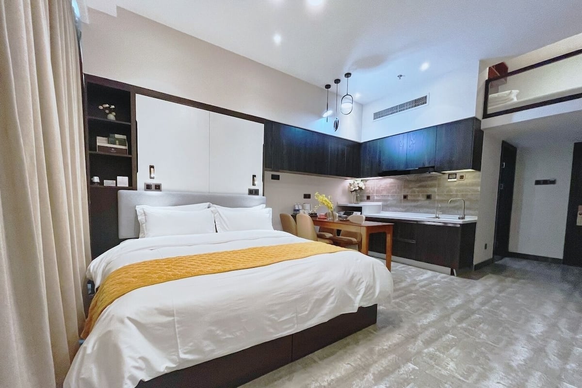 A modern hotel room with a large bed, a small desk, and a kitchenette.