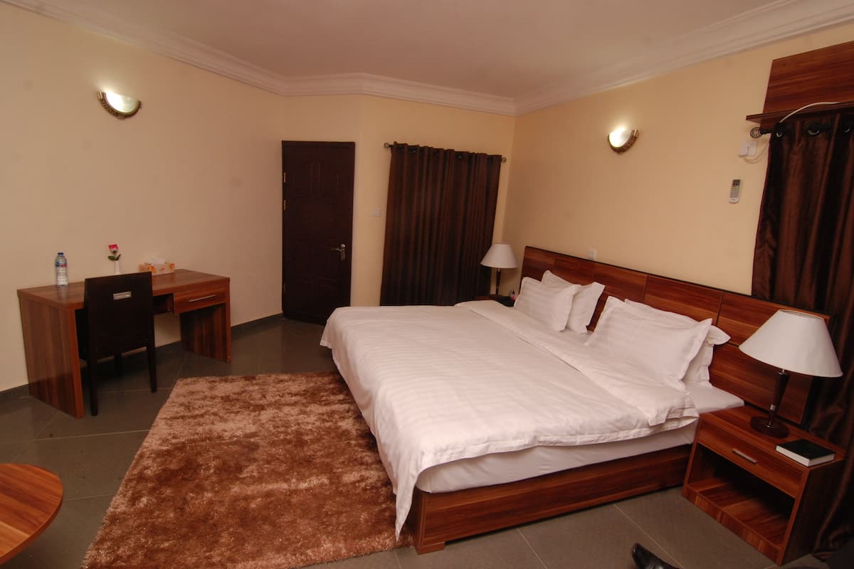Quarto de hotel com uma cama grande, uma escrivaninha com cadeira, uma mesinha de cabeceira de madeira, uma luminária e um tapete marrom.