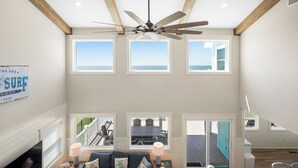 A spacious room with a ceiling fan, large windows, a view of the sea, and a sign on the wall. - Serenity by the Sea: Nostalgic Oceanfront Home with Room for the Whole Family (Oak Island)