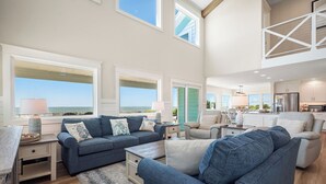 A spacious living area with large windows, a kitchen, and a balcony. - Serenity by the Sea: Nostalgic Oceanfront Home with Room for the Whole Family (Oak Island)
