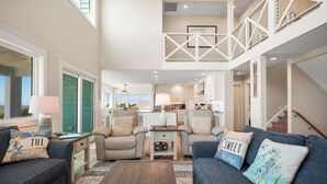 A spacious living area with a high ceiling, a staircase, and a kitchen in the background. - Serenity by the Sea: Nostalgic Oceanfront Home with Room for the Whole Family (Oak Island)