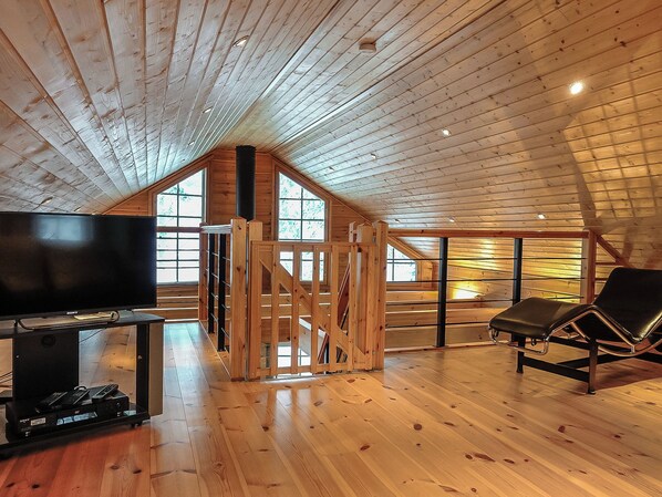 Une pièce mansardée spacieuse, avec un plancher en bois, une télévision et un fauteuil.