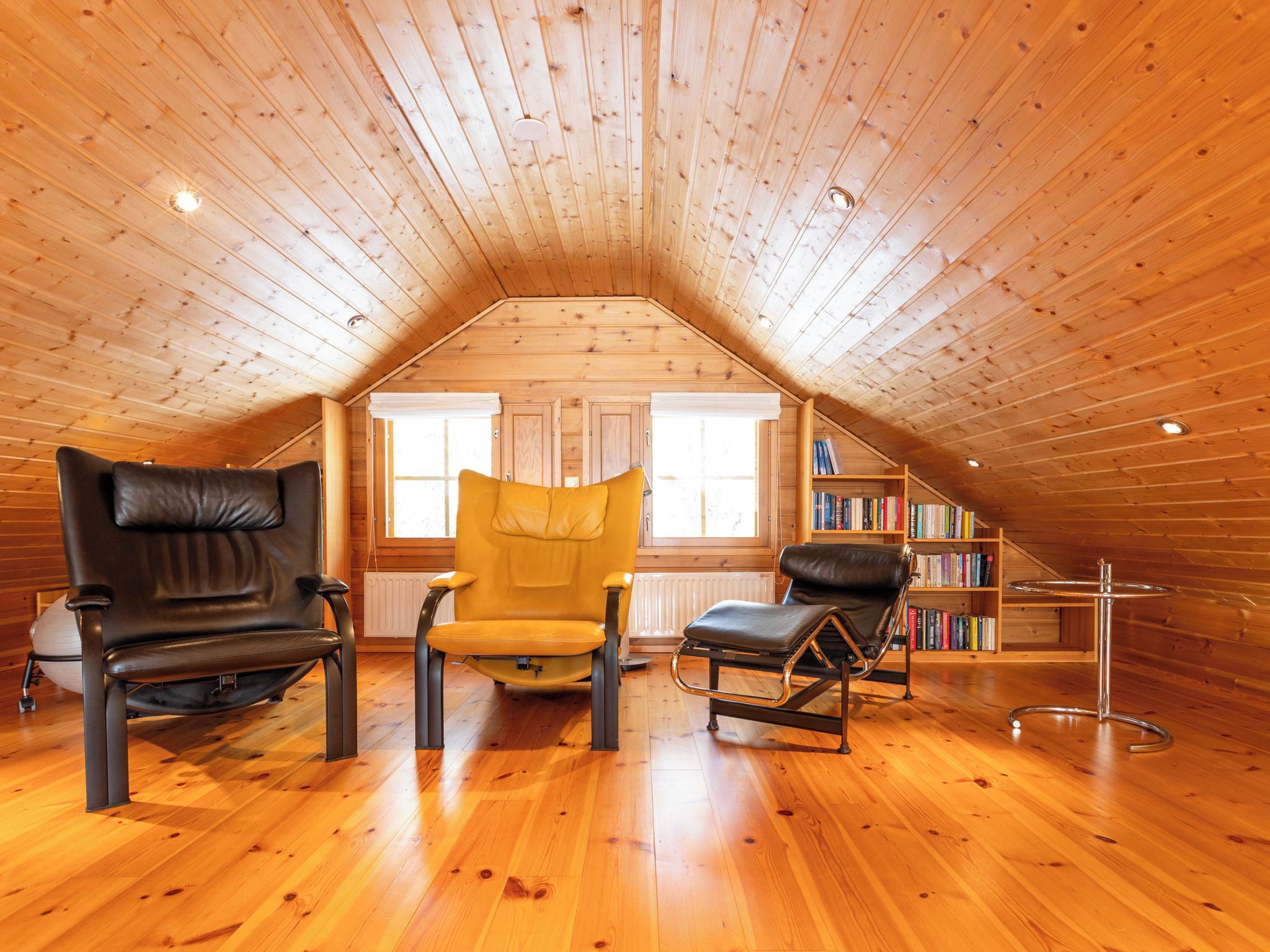 Quarto de sótão aconchegante, com ripas de madeira no teto, uma estante de livros e três poltronas de couro.