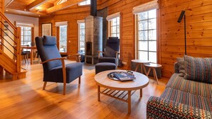 Interior de una cabaña de madera con chimenea de piedra, muebles de madera y ventanas amplias.