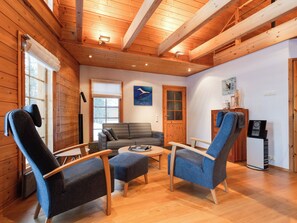 A cozy cabin interior with wooden walls, a sofa, two armchairs, a coffee table, and a fireplace.