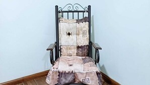 Una sedia con cuscino patchwork e struttura metallica, posizionata in un angolo su pavimento in legno e con una parete spoglia.