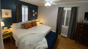 Une chambre à coucher comprenant un lit, un ventilateur de plafond, une table de chevet avec une lampe, une commode et une fenêtre avec des rideaux.