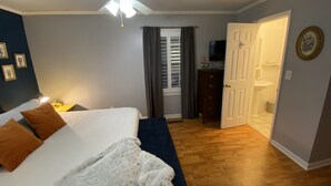 Une chambre à coucher comprenant un lit, un ventilateur de plafond, une table de chevet, une commode et une salle de bain visible à travers une porte ouverte.