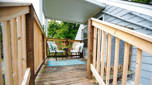 A wooden deck with a covered porch, two chairs, and a blue rug.