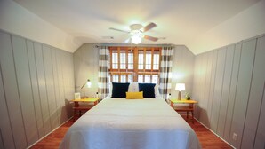 A bedroom with a large bed, a ceiling fan, two bedside tables with lamps, and a window with curtains.