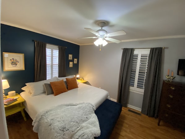 Un dormitorio con cama, ventilador de techo, mesita de noche con lámpara, cómoda y ventana con cortinas.