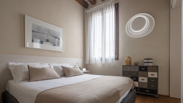 A bedroom with a bed, a nightstand, a dresser, a window with curtains, and a wall-mounted light fixture. - Santa Fosca Design Apartment R&R (Venecia)
