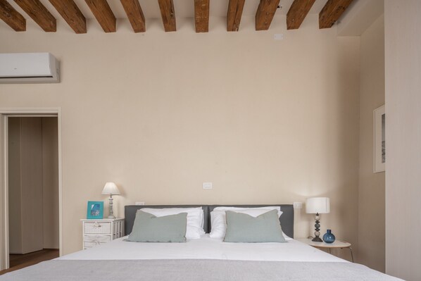 Un dormitorio con una cama grande, vigas de madera en el techo, mesitas de noche con lámparas y una puerta a la izquierda.