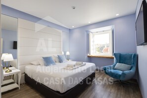 Une chambre moderne avec un grand lit, des tables de chevet, un fauteuil bleu et une fenêtre donnant sur un beau paysage.