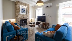 A modern living room with a stone fireplace, blue sofas, a flat-screen TV, and a large window. - Villa S. Lourenço by ACasaDasCasas (Ericeira)