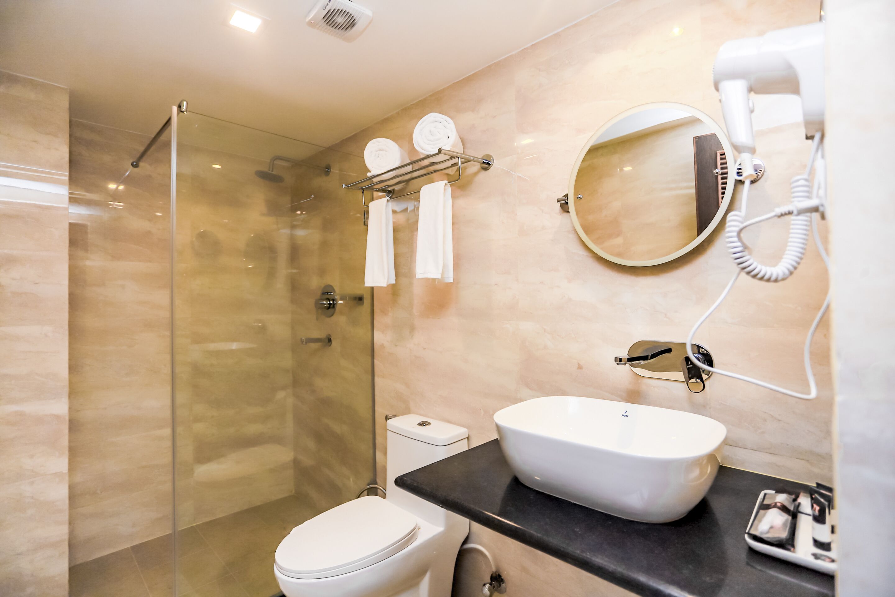 Ein modernes Badezimmer mit einer gläsernen Duschkabine, einer weißen Toilette, einer schwarzen Ablage mit einem weißen Waschbecken und einem an der Wand befestigten Föhn.