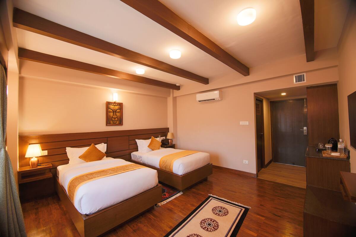 A hotel room with two beds, wooden beams on the ceiling, a nightstand with a lamp, a painting on the wall, and a door leading to another room.