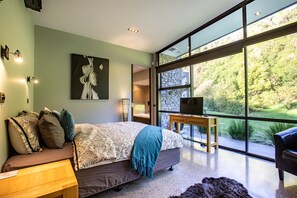 Un dormitorio moderno con una cama grande, una mesita de noche de madera, un televisor y vistas al exterior a través de amplias ventanas.