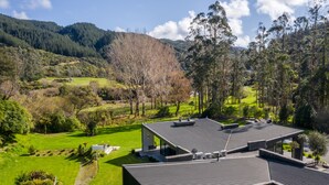 Une maison moderne dotée d’un toit en pente, entourée de champs verdoyants et d’arbres, avec une montagne en toile de fond.