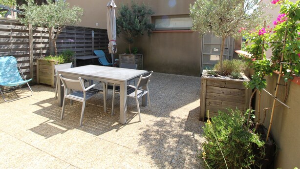 Une terrasse extérieure avec une table et des chaises, entourée de plantes et d’une clôture en bois.