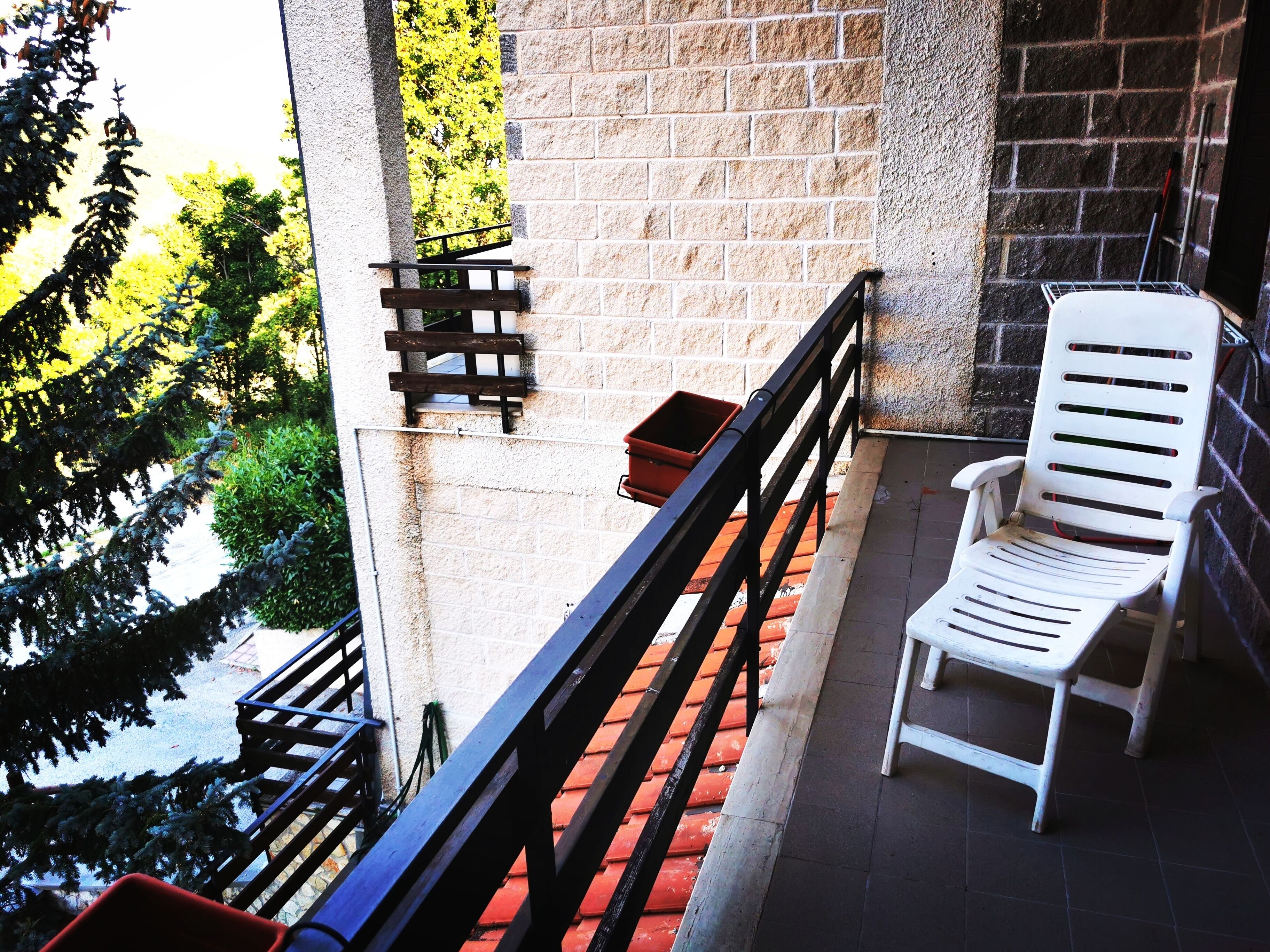 Un balcone con una sedia di plastica bianca, una piccola panca di legno e un vaso rosso.