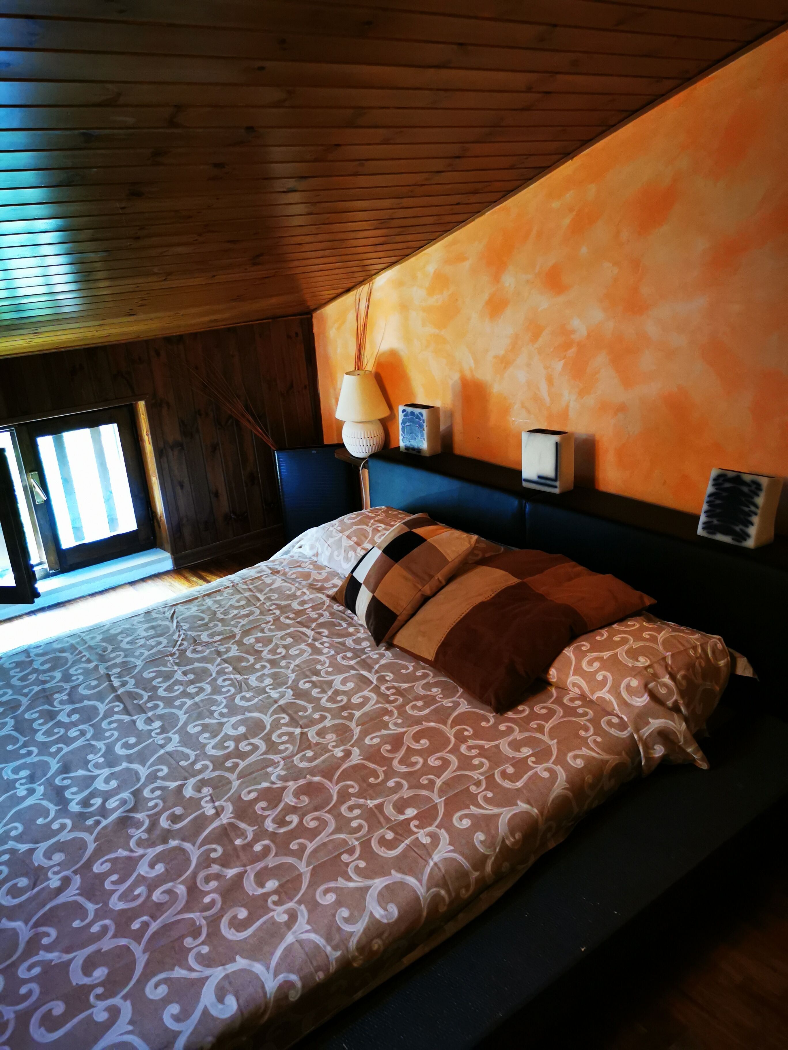 A bedroom with a wooden ceiling, a bed with patterned bedding, a nightstand with a lamp, and a window with blinds.